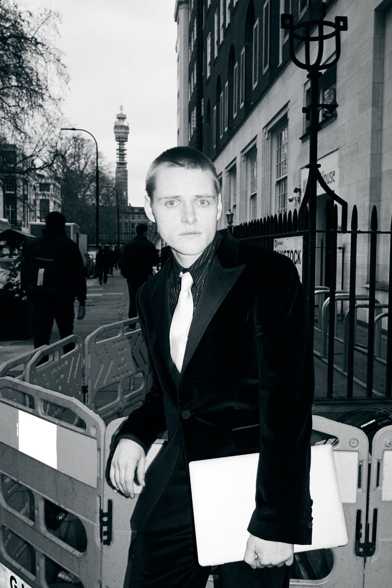 Monochrome portrait of a slender adult with a light complexion in a dark suit on a London street, leaning against a barrier.