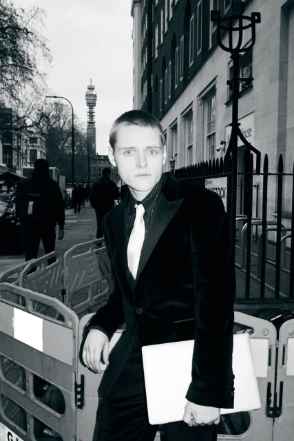 Monochrome portrait of a slender adult with a light complexion in a dark suit on a London street, leaning against a barrier.