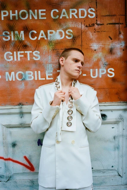 Person in an all-white suit standing in front of a wall with graffiti. The suit is accessorized with jewelry. 