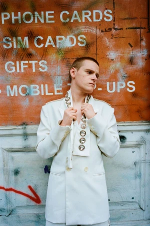 Person in an all-white suit standing in front of a wall with graffiti. The suit is accessorized with jewelry. 