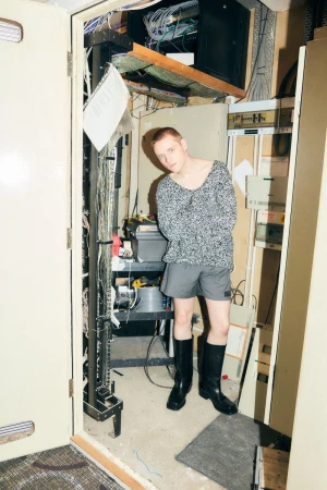 Young man poses in a cluttered room with equipment, wearing a grey sweater, shorts, and black boots, standing near an open doorway.