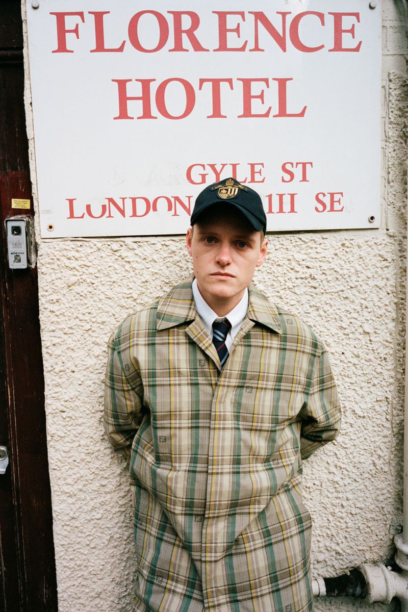 Young adult poses outdoors in a plaid coat, baseball cap, and tie near a hotel sign on a textured building exterior in daylight.