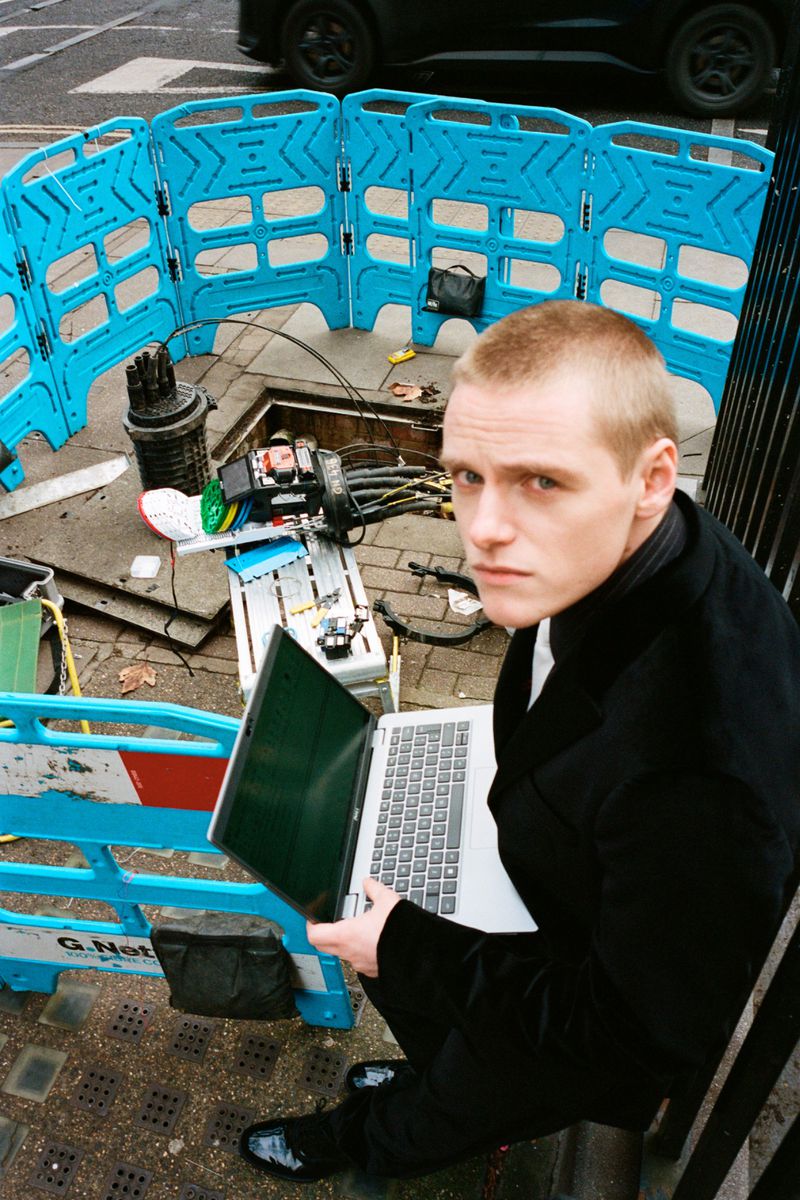 Adult male uses laptop next to a utility access point. Blue safety fencing surrounds the area. A car is parked in the background.