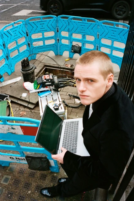 Adult male uses laptop next to a utility access point. Blue safety fencing surrounds the area. A car is parked in the background.