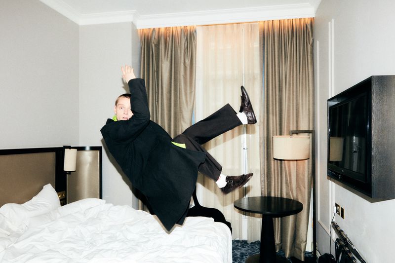 A man in a black coat mid-air on a bed in a hotel room with a side table and a television in the background, creating a dynamic scene.