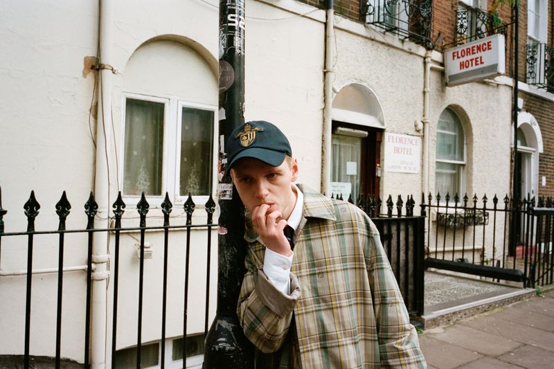 Portrait of a young adult man in a baseball cap and plaid jacket, standing near a black fence and a light pole in an urban setting.
