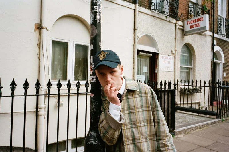Portrait of a young adult man in a baseball cap and plaid jacket, standing near a black fence and a light pole in an urban setting.