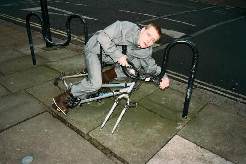 Man in a grey suit poses on top of a white bicycle in an urban environment with black metal bike racks on a city street.