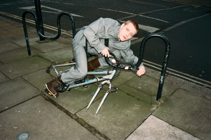 Man in a grey suit poses on top of a white bicycle in an urban environment with black metal bike racks on a city street.