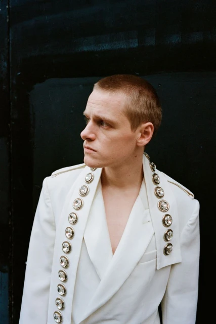 Stylish portrait of a fair-skinned adult male wearing a white jacket with jeweled embellishments against a dark background.