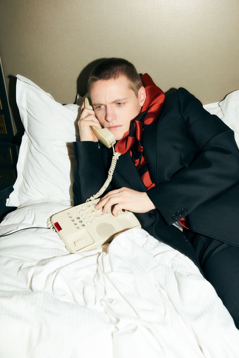 Adult male in black suit jacket and patterned scarf poses with a vintage telephone while reclining on a white, rumpled bed in a room.