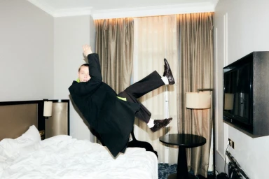 Man wearing a black coat mid-air falling backwards into a hotel bed for Behind the Blinds photographed by Oliver Webb.