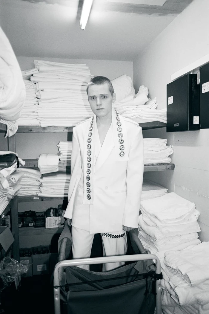 Young man in a white suit standing in a narrow storage room filled with stacked white linens for Behind the Blinds.