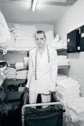 Young man in a white suit standing in a narrow storage room filled with stacked white linens for Behind the Blinds.