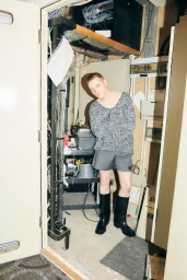 Lewis Gribben in a textured grey knit sweater, grey shorts, and tall black boots standing inside a utility or server room.