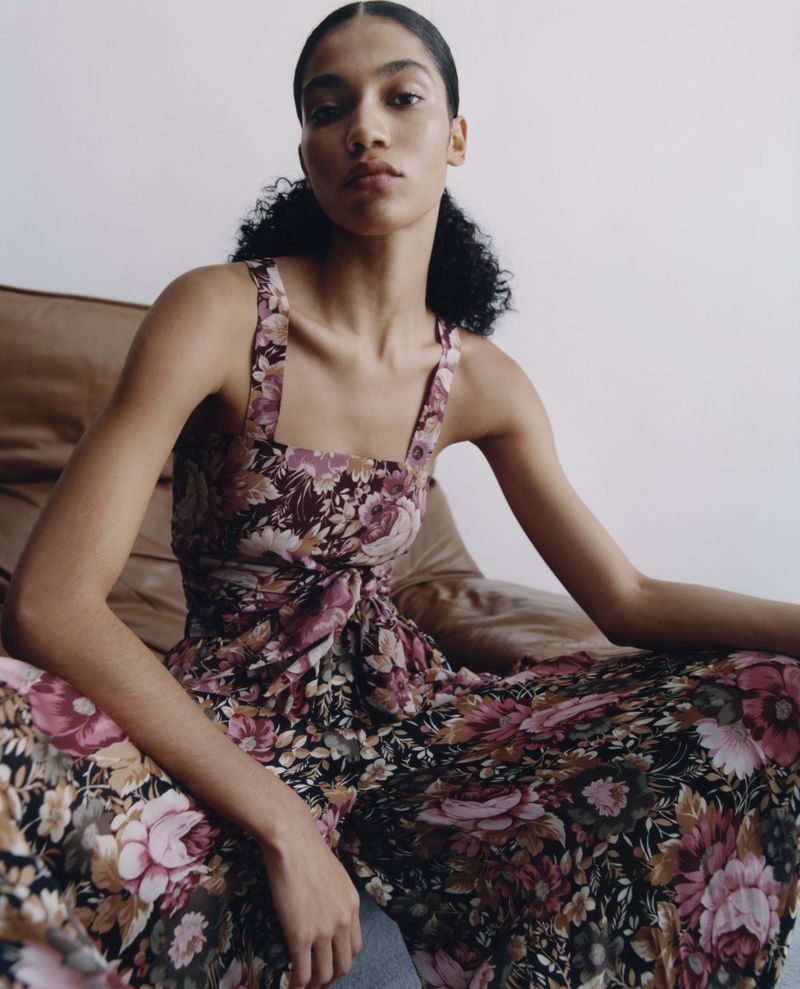 An adult female model sits indoors wearing a floral print dress on a brown chair in a studio setting against a neutral background.