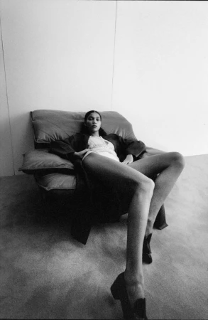 A slender woman rests in an armchair wearing a coat and heels, black and white studio shot, minimalist composition.