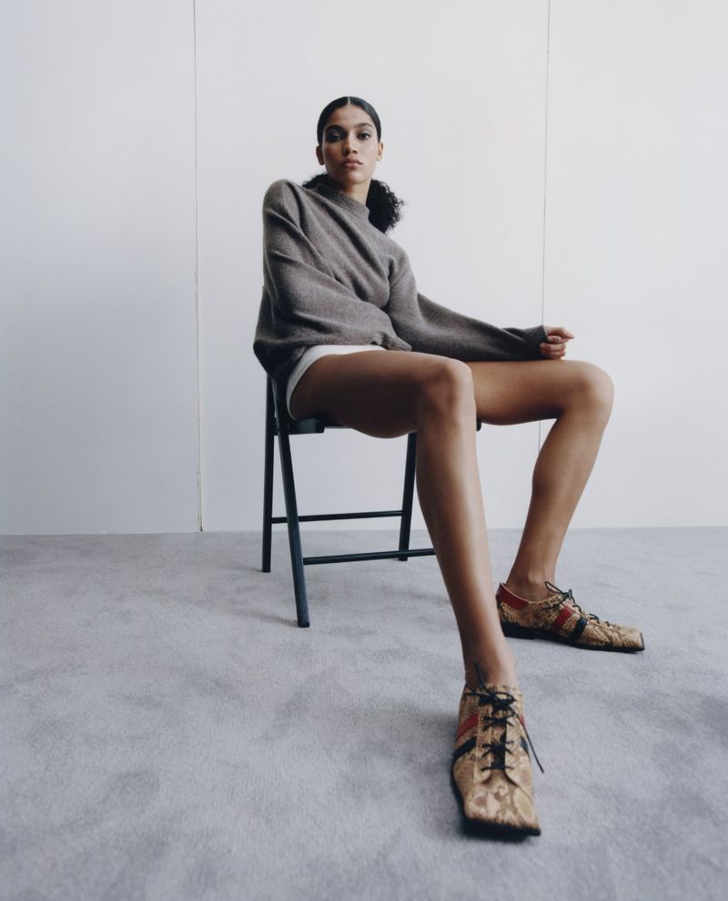 Model Raynara Negrine sitting on a black folding chair against a neutral grey studio background, wearing a taupe sweater and patterned lace-up shoes.