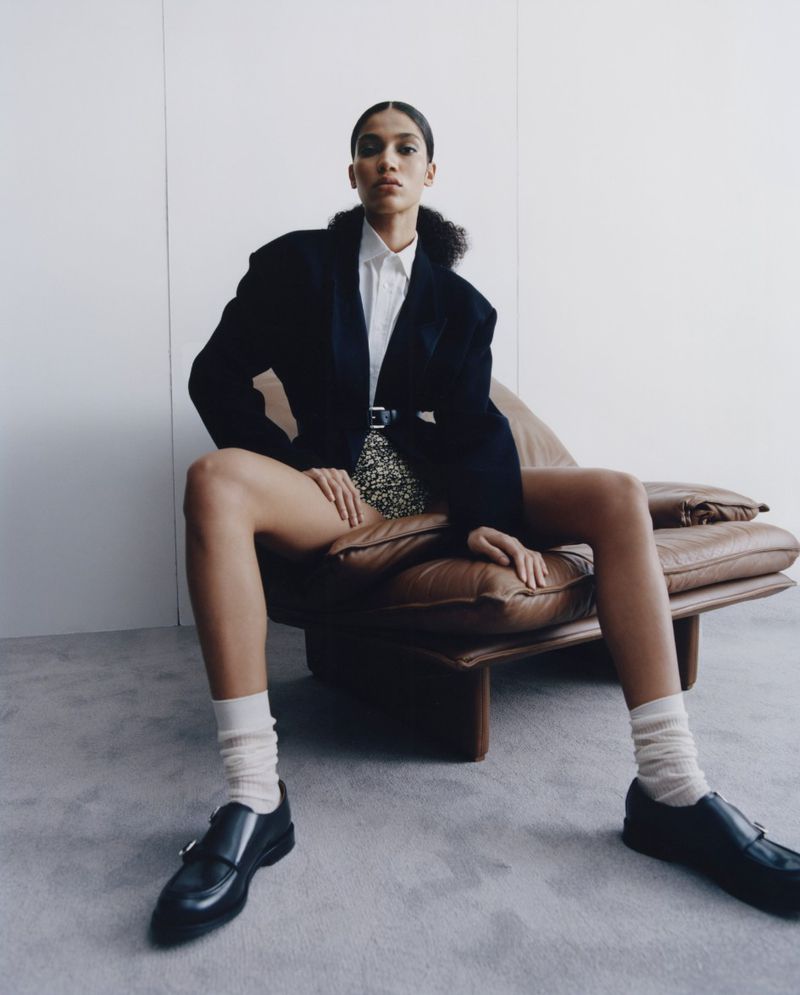 Model seated on a brown leather armchair wearing a black blazer, white shirt, and loafers against a plain wall.