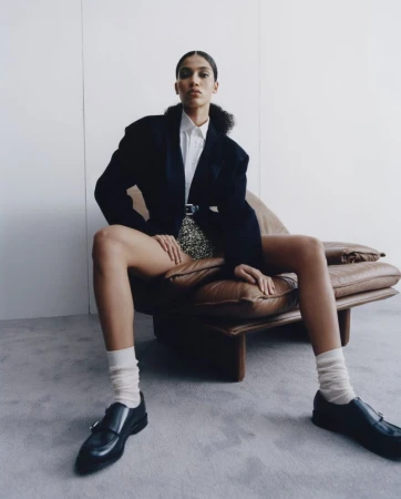 Model seated on a brown leather armchair wearing a black blazer, white shirt, and loafers against a plain wall.