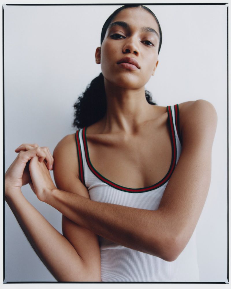 Portrait of a young adult woman with dark hair in a ponytail, wearing a white ribbed tank top, arms folded, against a white wall.