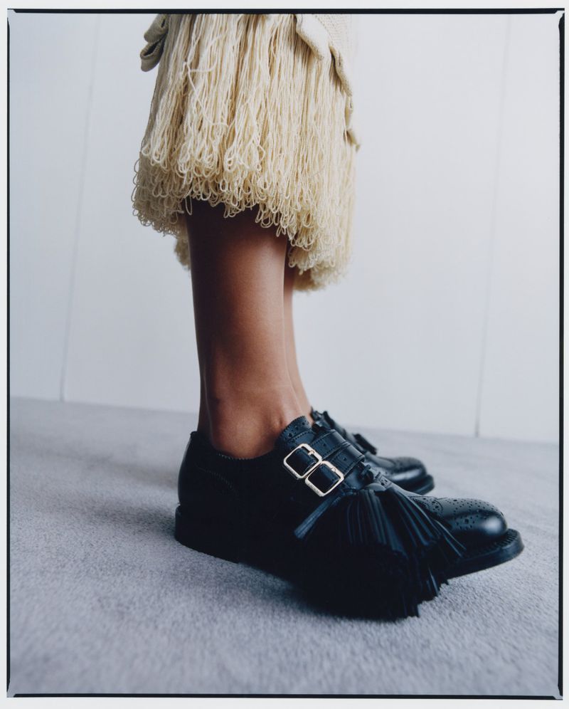 Low angle shot of a woman wearing a cream fringe skirt and black leather shoes with tassels against a neutral background.