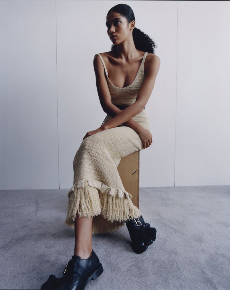 A slender African-American woman sits on a box in a sleeveless knitted top and matching skirt with black shoes in a minimalist indoor setting.