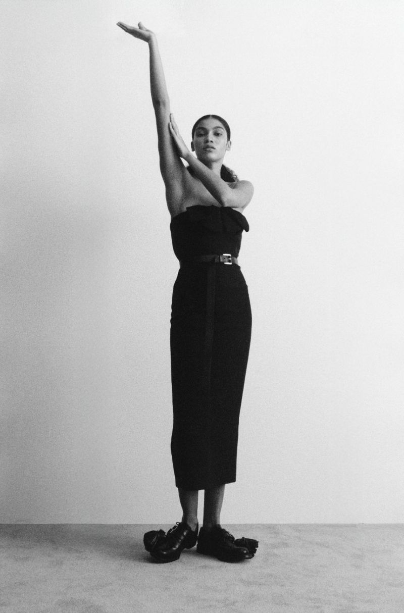 Full-length shot of a woman in a black strapless dress and sculptural shoes, with arms raised in a minimalist studio setting.
