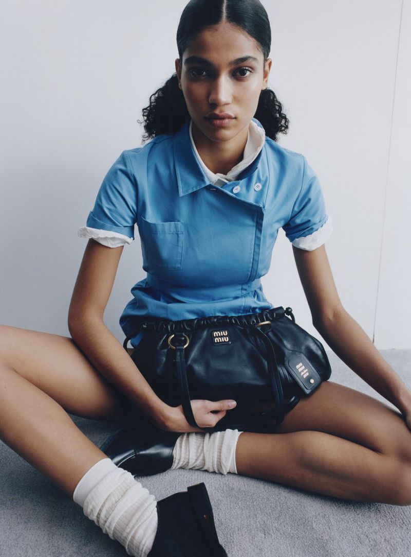 Model Raynara Negrine sitting cross-legged wearing a blue uniform dress holding a black leather handbag against a plain background