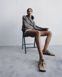 Model Raynara Negrine sitting on a black folding chair against a neutral grey studio background, wearing a taupe sweater and patterned lace-up shoes.