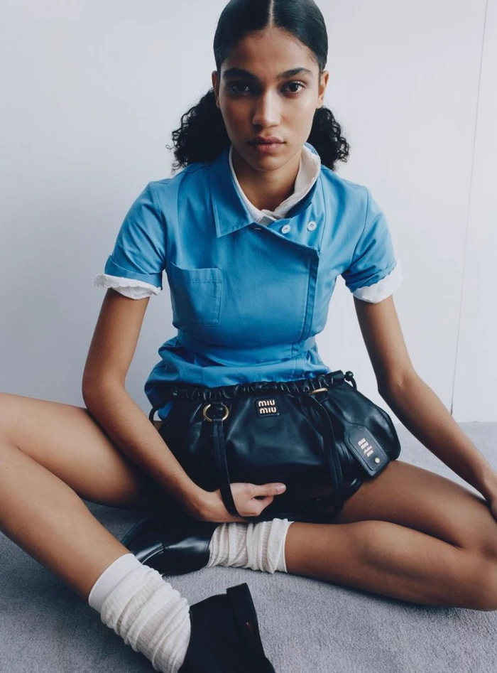 Model Raynara Negrine sitting cross-legged wearing a blue uniform dress holding a black leather handbag against a plain background