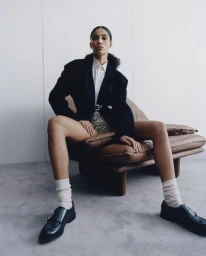 Model seated on a brown leather armchair wearing a black blazer, white shirt, and loafers against a plain wall.