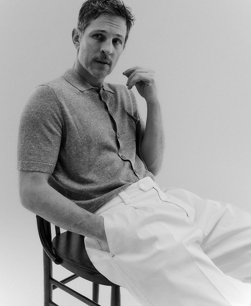 Black and white portrait of a male model with dark hair, wearing a grey polo shirt and white pants, sitting on a chair in a studio.