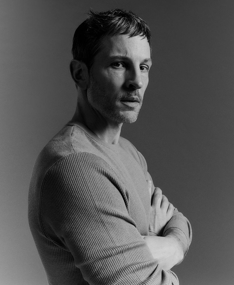 Monochrome portrait of an adult male with arms crossed, wearing a textured sweater against a neutral grey background, medium shot.
