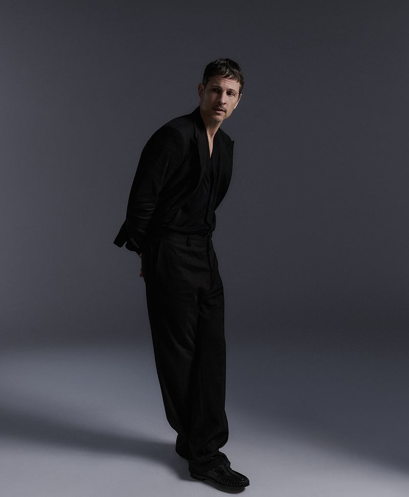 Studio shot of an adult male in a black suit, posed in a three-quarter view against a neutral gray background with soft, even lighting.