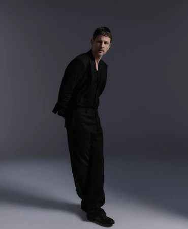 Studio shot of an adult male in a black suit, posed in a three-quarter view against a neutral gray background with soft, even lighting.