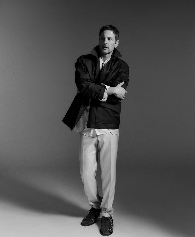 Full shot of adult male in dark jacket, white shirt, light pants, and dark sandals, arms crossed, in a neutral grey photo studio.