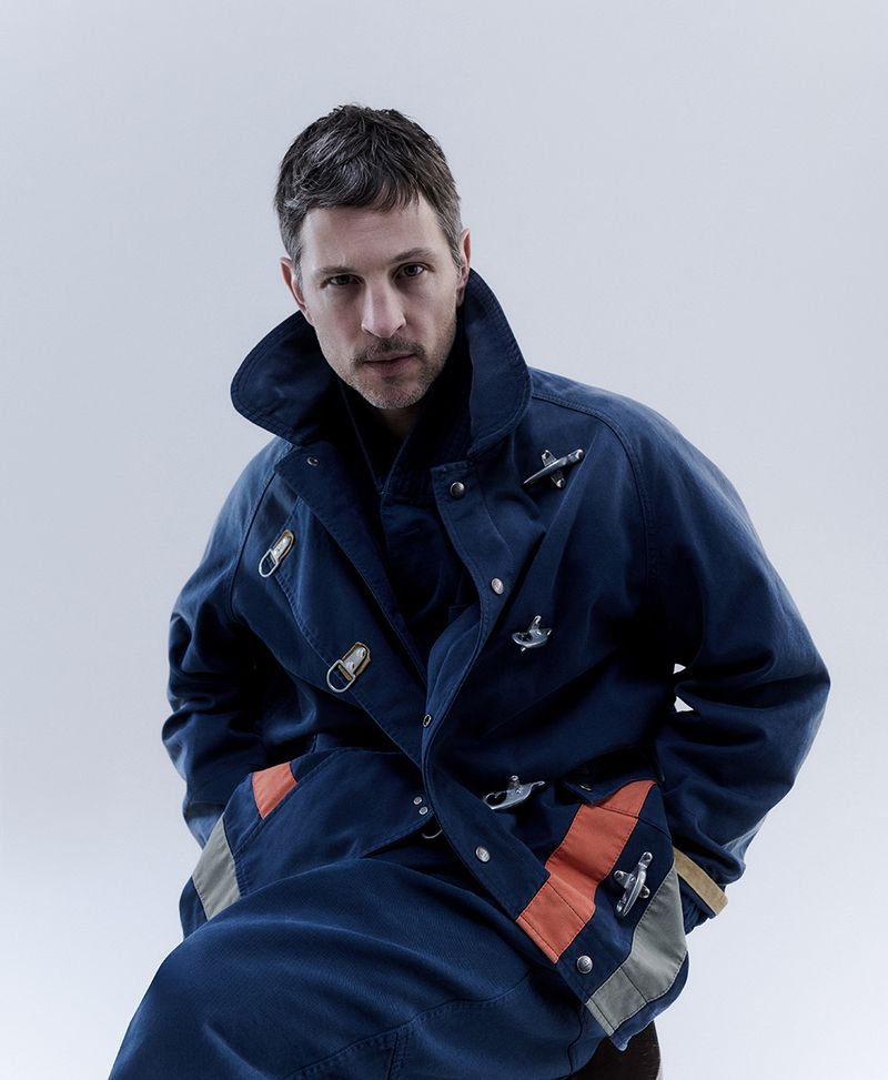 Portrait of an adult male wearing a navy-blue coat with orange and grey accents posing in a photo studio with a neutral background.