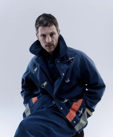 Portrait of an adult male wearing a navy-blue coat with orange and grey accents posing in a photo studio with a neutral background.