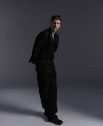 Man wearing a black suit jacket and trousers posing against a dark grey studio background for Style Magazine Italy fashion issue.