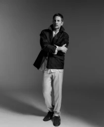 Monochrome fashion portrait of a man in a dark jacket and light trousers standing against a seamless grey backdrop.
