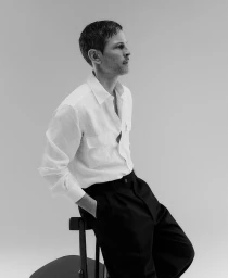 Man in white button-down shirt and black trousers sitting on a stool against a neutral background
