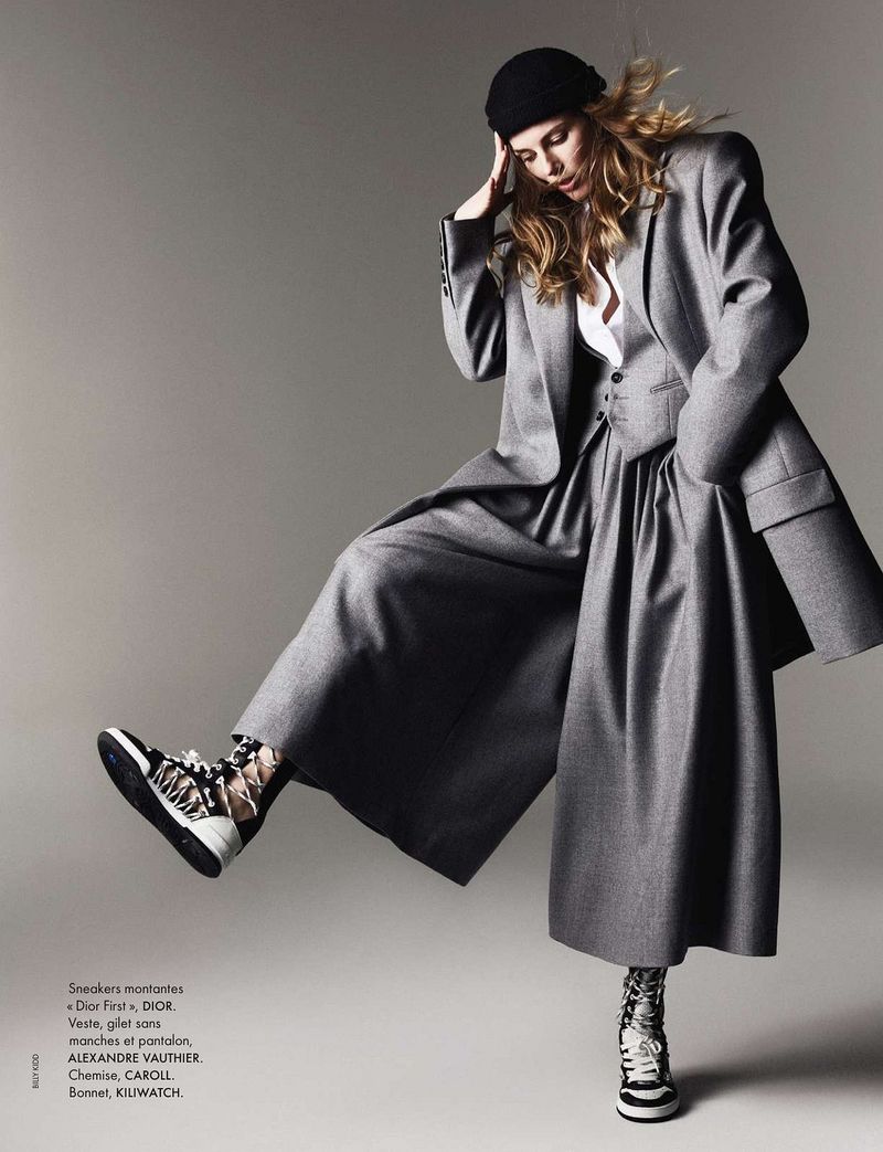 Fashion editorial shot: a woman in a grey suit and beanie hat poses with one leg raised, showcasing patterned sneakers, against a grey background.
