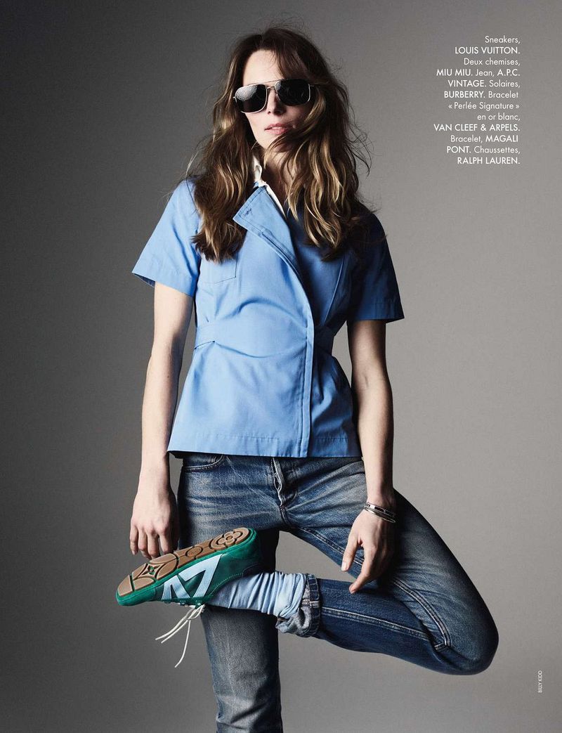 Fashion editorial shot of a woman posing in a blue top and jeans, wearing sunglasses and shoes, set against a grey background.