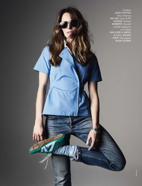 Fashion editorial shot of a woman posing in a blue top and jeans, wearing sunglasses and shoes, set against a grey background.