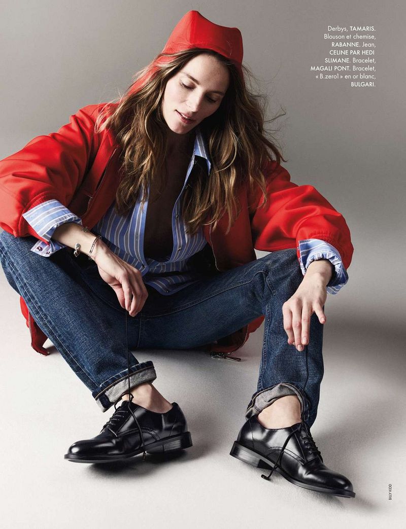 Editorial fashion shot of a woman in a red jacket, blue jeans, and black shoes, sitting on a plain white surface.