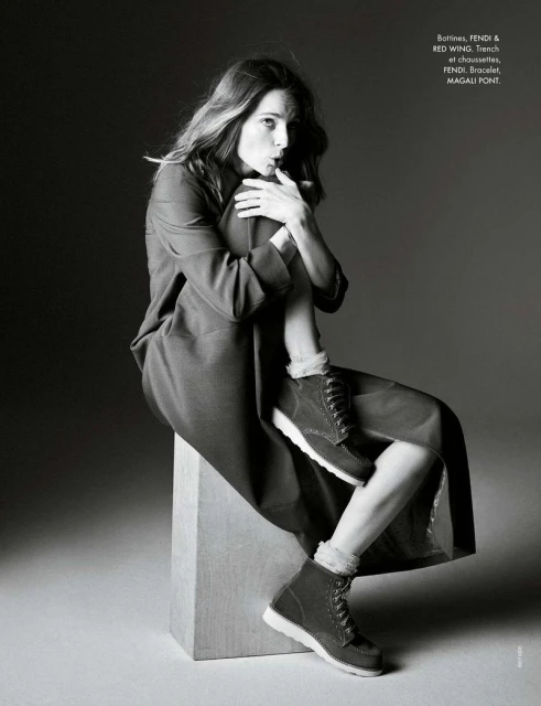 Black and white portrait of a woman in a jacket and boots, sitting on a block, posing in a photo studio, fashion editorial shot.