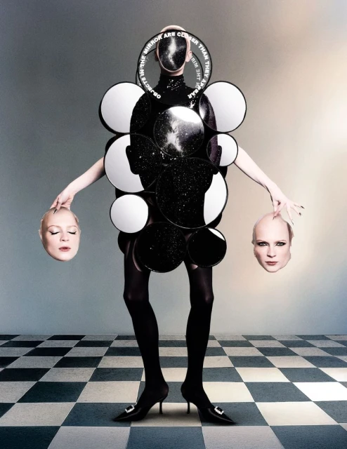 Avant-garde fashion shot featuring a person in a mirror-decorated garment holding two mannequin heads on a checkered floor.