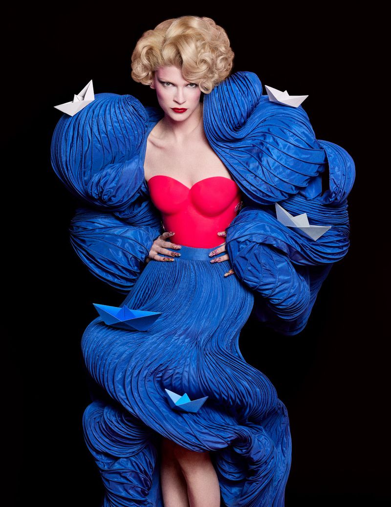 Fashion model in blue sculptural dress with paper boat details against a black backdrop, wearing red top and styled blonde hair.