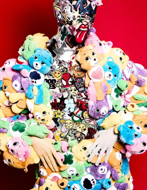 Person covered in plush Care Bears and sticker-covered fabric against a red background, with hands visible in sheer gloves.